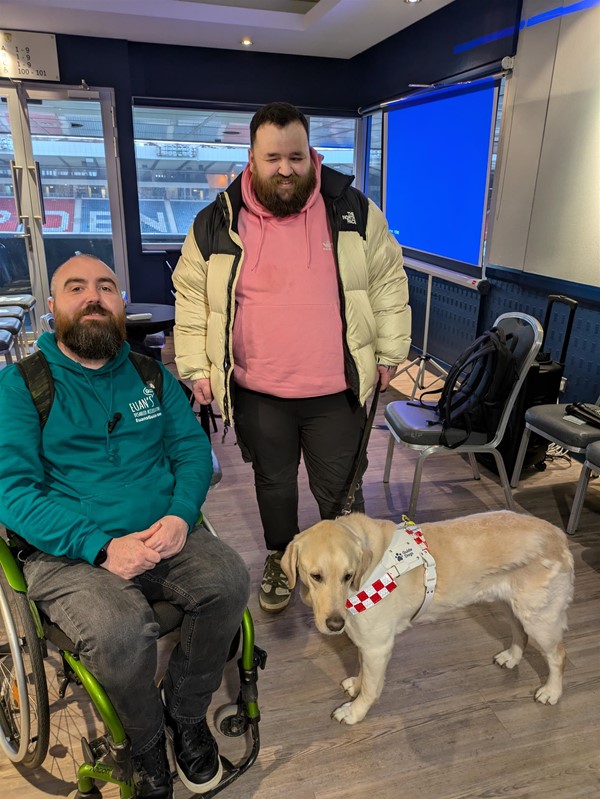 Image of Joe, Jon and Sam the Guide Dog. Joe is a wheelchair user with a shaved head and brown beard, Jon stands beside him and has dark hair and a dark beard. Sam is a blonde labrador.