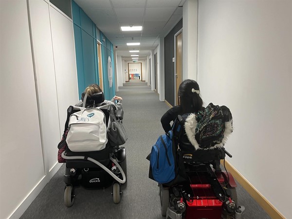 Two power chair users going down the hallway past the double doors.