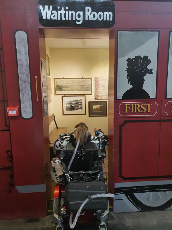 Image of Claire going into waiting room carriage area to show the width of the entrance compared to her wheelchair.