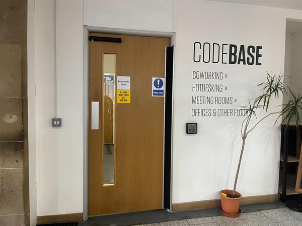 Wooden automatic door with a glass window pane down the side. There is a silver square button placed on the white wall to the right of the door.