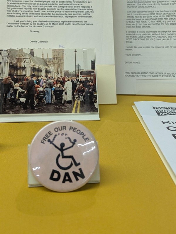 Image of a pink badge showing a wheelchair user symbol, with arms breaking a chain. It reads "Free our People" and "DAN" at the bottom.