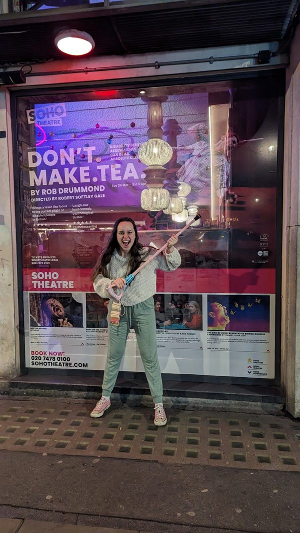 ID: Katie, the writer of this review, is standing in front of the theatre. In the window behind her is a poster for the production that reads ' Don't. Make. Tea' and then lists some of the cast and creatives. She is holding her Walksit from Glamsticks (a mobility aid brand) as if she is about to hit someone with it - this is a reference to a plot point in the show. Katie is a young white woman, with long brown hair hanging over her cream jumper, which compliments her bloat-proof green trousers. She is wearing pink converse the same colour as her stick.