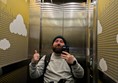 Image of  Joe reflected in the lift's mirror. He's a white man with a beard, wearing a blue hat an grey jumper