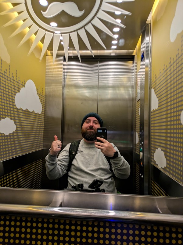 Image of  Joe reflected in the lift's mirror. He's a white man with a beard, wearing a blue hat an grey jumper