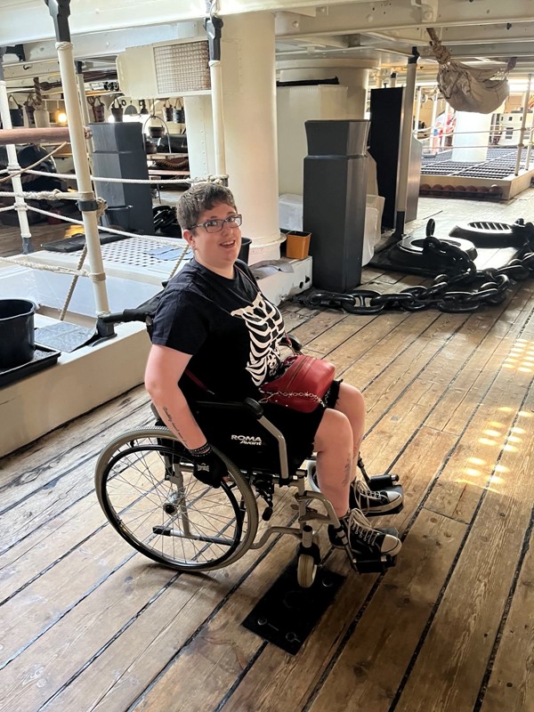 I am a white, non-binary person with short, dark blonde hair and multiple tattoos. I am sat in a self-propelled manual wheelchair on the gun deck of the HMS Warrior, surrounded by cannons, ropes, and hammocks.