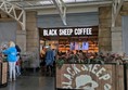 Exterior of Black Sheep Coffee. There is low fencing with the black sheep logo. Illuminated lettering that reads BLACK SHEEP COFFEE is above the door.