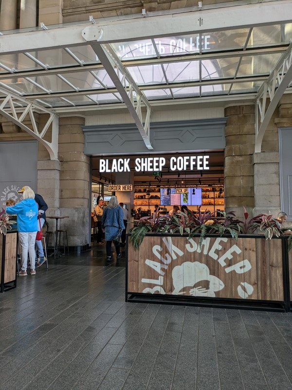 Exterior of Black Sheep Coffee. There is low fencing with the black sheep logo. Illuminated lettering that reads BLACK SHEEP COFFEE is above the door.