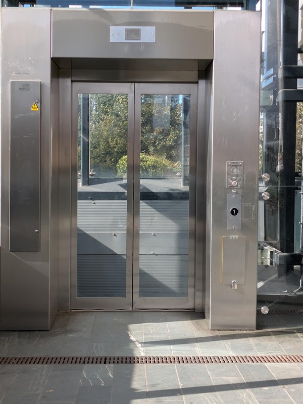 Image of the lift access, a metal booth with sliding doors. To the right is the call button