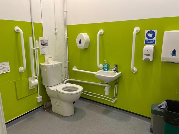 Spacious toilet with a small black bin to the right of the image. There is a grab rail that has been raised against the wall and a red cord hanging freely to the left of the image. Soap and paper towel dispensers are installed on the wall.