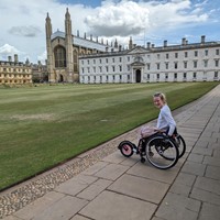 roz, a white person with blonde hair sits in their quickie argon wheelchair with a pink freewheel attachment. They are infront of Kings College Cambridge, looking at beautifully manicured lawns and the iconic chapel