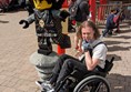 Logan (a white man with shoulder length brown hair and black dungarees) does a wheelie in his wheelchair next to a life sized Lego Ninjago model.