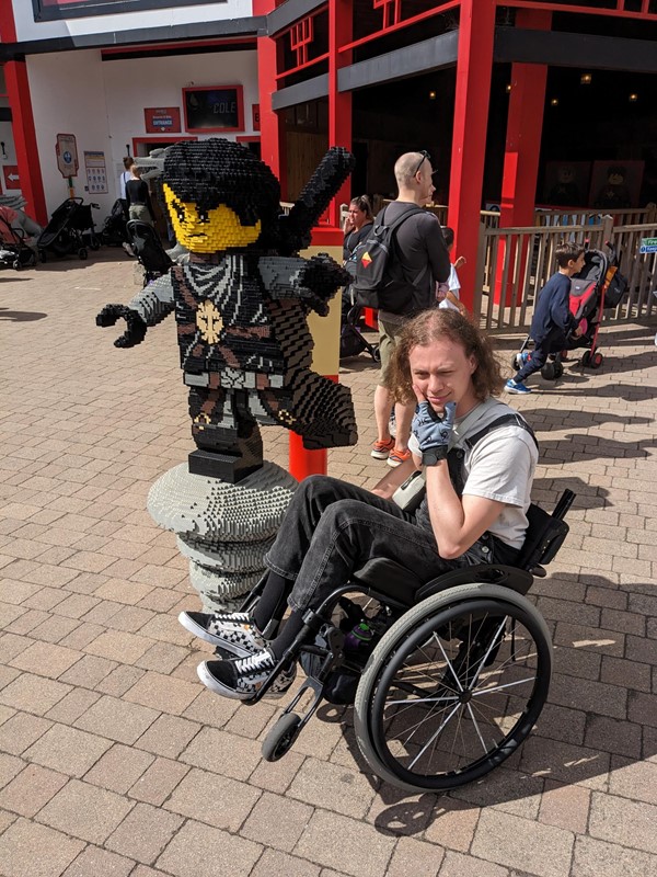 Logan (a white man with shoulder length brown hair and black dungarees) does a wheelie in his wheelchair next to a life sized Lego Ninjago model.