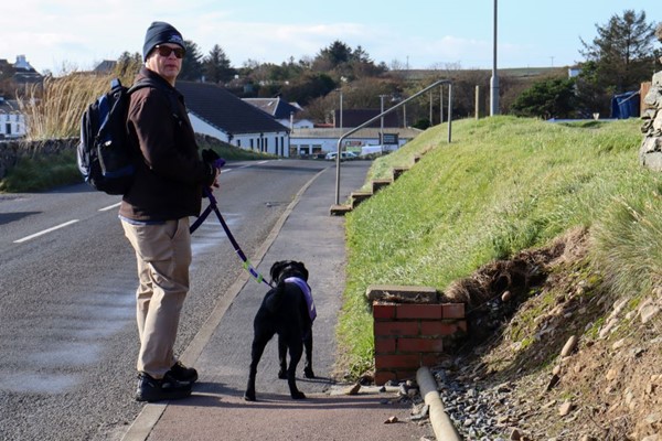 The pavement continues with no dropped kerbs and there is a flight of steps which makes the pavement too narrow for a wheelchair to pass. Man with dog looks back with a concerned expression.