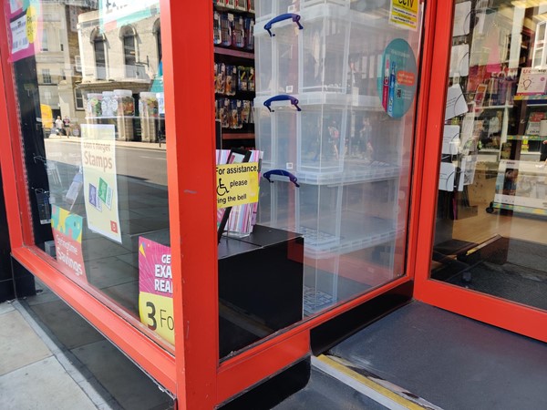 The entrance to the shop with a sign showing a wheelchair symbol and the words 'For assistance please ring the bell'. The doorbell button unit is missing.