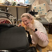 roz, a white person with blonde hair, sits on the floor in front of a quickie helium wheelchair. they hold a spanner in one hand and an allen key in the other, and are grinning at the camera