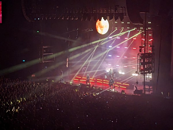 Ovo stage with red lighting and search lights going out over the heads of a full standing area of fans.

Fall Out Boy are on the stage plating instruments.