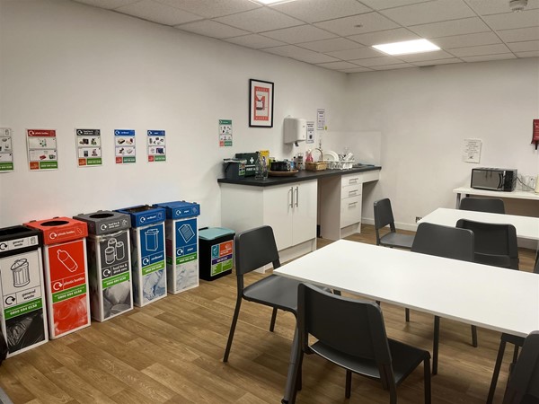 Employee kitchen with 2 rows of tables, 5 bins of various types and a counter to the end of the wall. There is also a microwave and a sink.