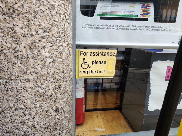 Concealed behind a pillar, a sign with a wheelchair symbol on it, the words FOR ASSISTANCE PLEASE RING THE BELL, and a worn-out spot where the doorbell button used to be but is no longer there.