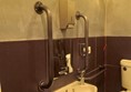 a photo of the sink in the bathroom. it shows a low sink, a small mirror above it and a metal soap dispenser above the mirror. metal grab bars surround the mirror.