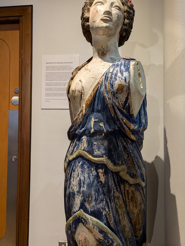 Image of a figurehead, a distressed wooden sculpture of a woman with brown hair in a blue dress, the arms are absent