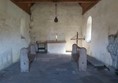 Image of a very simple interior of a stone built chapel. Stone flooring. The alter is a plain wooden beam, there is a small row of church candles next to it. There is a crucifix in the corner possibly made of driftwood.