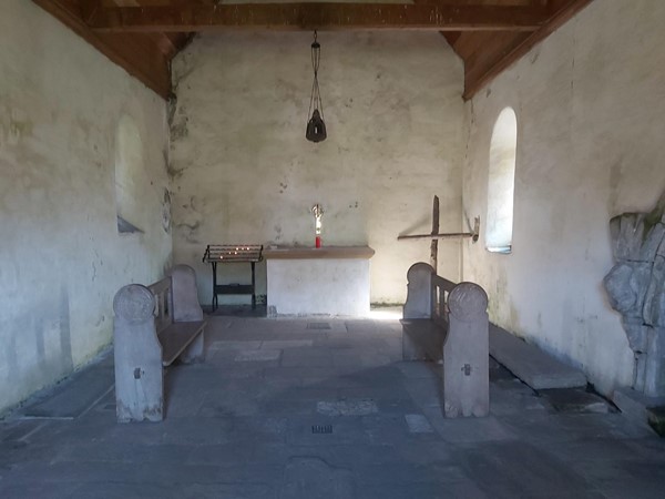 Image of a very simple interior of a stone built chapel. Stone flooring. The alter is a plain wooden beam, there is a small row of church candles next to it. There is a crucifix in the corner possibly made of driftwood.