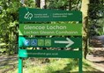 A green sign with directions for Glencoe Lochan, including icons for parking, wheelchair access, picnic area, and walking trail. The sign features text in both English and Gaelic.