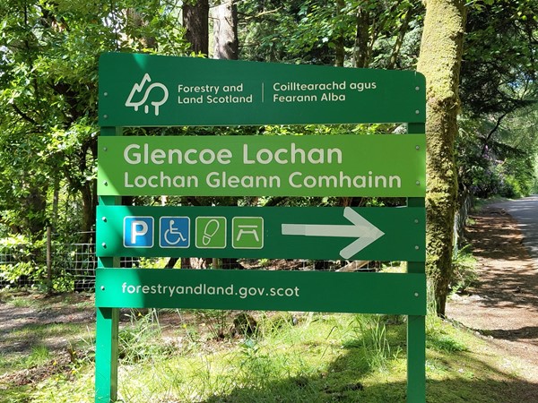 A green sign with directions for Glencoe Lochan, including icons for parking, wheelchair access, picnic area, and walking trail. The sign features text in both English and Gaelic.
