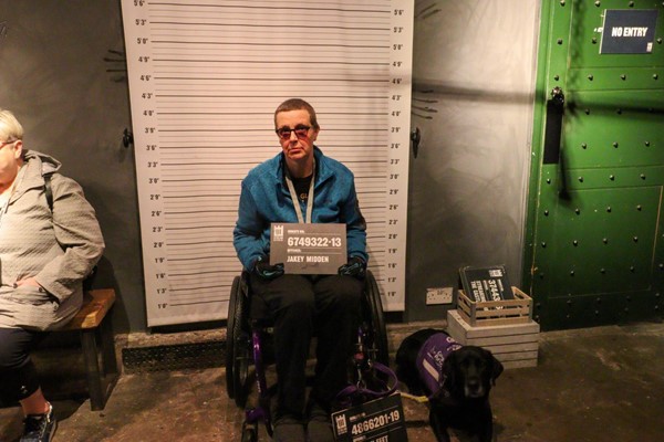 Lady in wheelchair with assistance dog, sitting in front of height chart. The lady and the dog are both holding prison number cards.