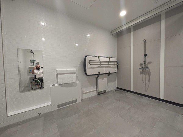 Image of the toilet, showing the change bed, shower, large towel dispenser and mirror. The photographer, a bearded white man sits in a wheelchair giving a thumbs up to the mirror.