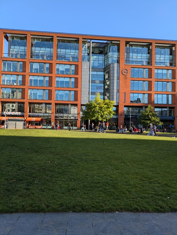 Image of a grassy area, the building at the back features a Popeyes and Ask Italian restaurant